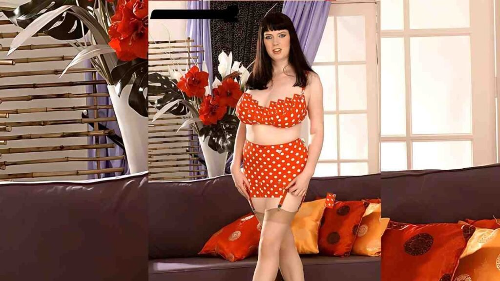 Emily Cartwright in red polka-dot with garter stockings poses in a living room with a sofa and orange cushions.