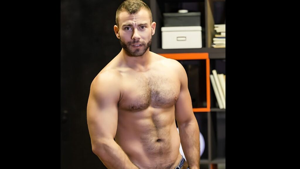 Enzo Rimenez with a short beard stands in gym room, flexing as orange-framed shelves and boxes loom behind him.
