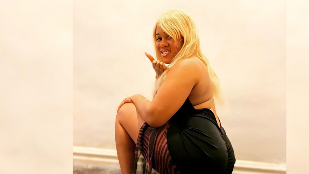 Gemini Lovell in a black backless dress crouches with one knee up, blowing a kiss toward the camera in a bright studio.