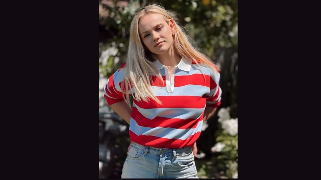 Ginta Lapiņa with long blonde hair poses outdoors in a red, white, and blue striped rugby shirt and jeans, looking calmly at the camera.