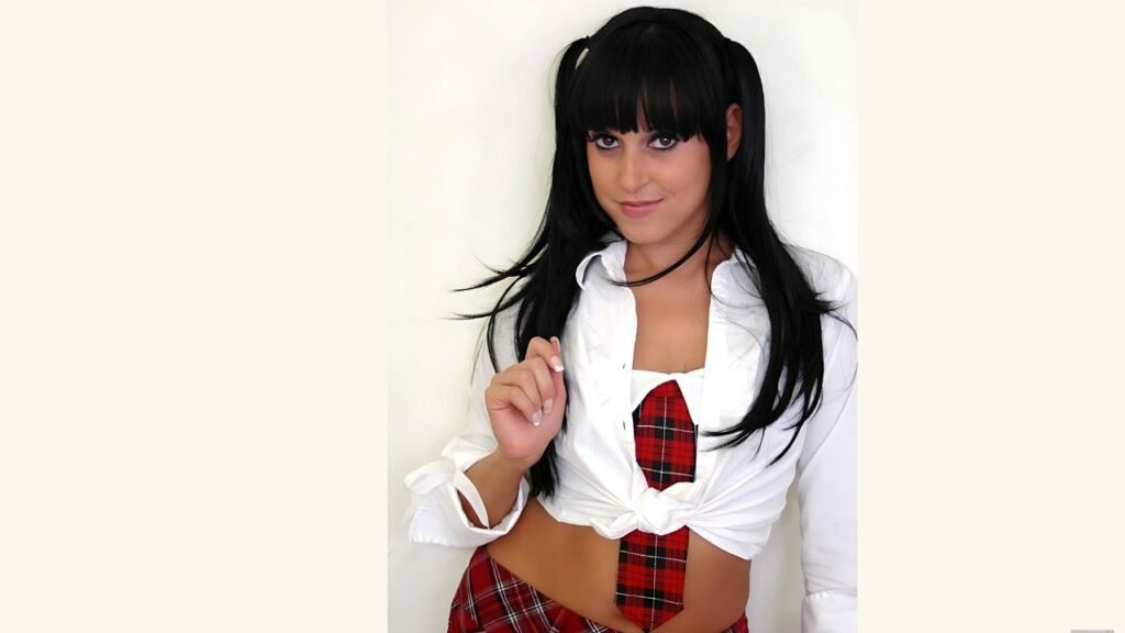 Giselle Vega with black twin ponytails and bangs wears a tied white shirt over a red plaid schoolgirl uniform, posing.