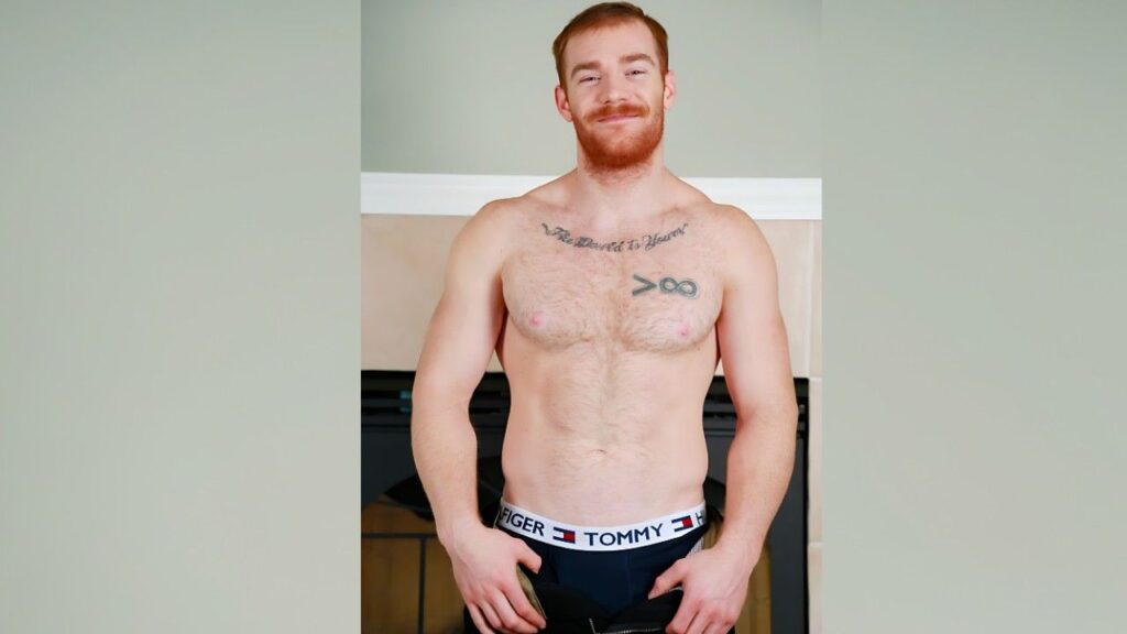James Jamesson with red hair and a beard stands in front of a fireplace, showing chest tattoos and waistband of his underwear.