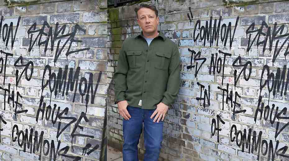 Jamie Oliver in an olive green jacket and blue jeans stands before a graffiti-covered brick wall, looking at the camera.