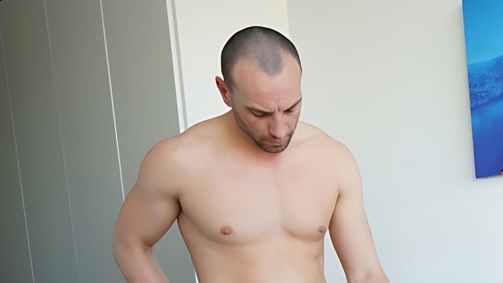 Jayce Hardy with a shaved head looks down in a room, pale walls, left sliding closet doors, and blue artwork on the right.