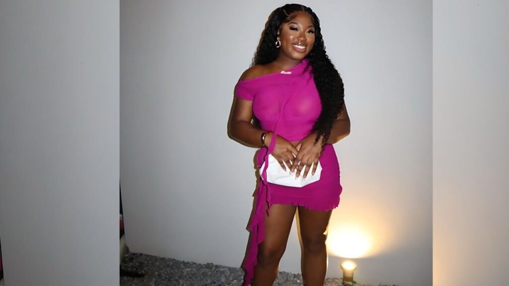 Jennifer B in magenta off-shoulder mini dress with fringe hem, holding a white clutch, standing by a warm-lit wall.