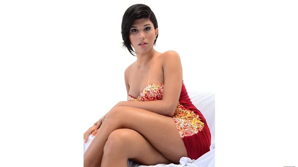 Jessica Versace in a red strapless dress with gold sequins sits cross-legged on white fabric, gazing at the camera.