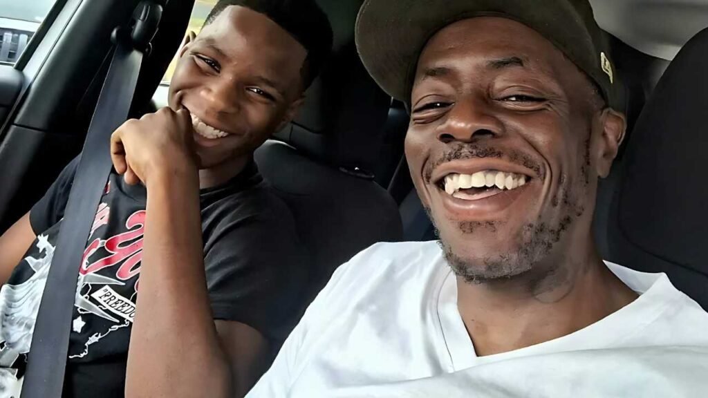 Jett Black in a car take a selfie, smiling; one wears white and a cap, the other a black graphic tee, both with seat belts.