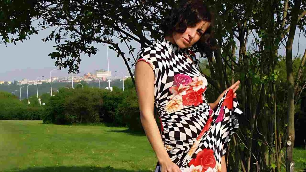 Joan White in a bold black-and-white checkered dress with pink rose prints tilts her head in a sunny park beside trees.