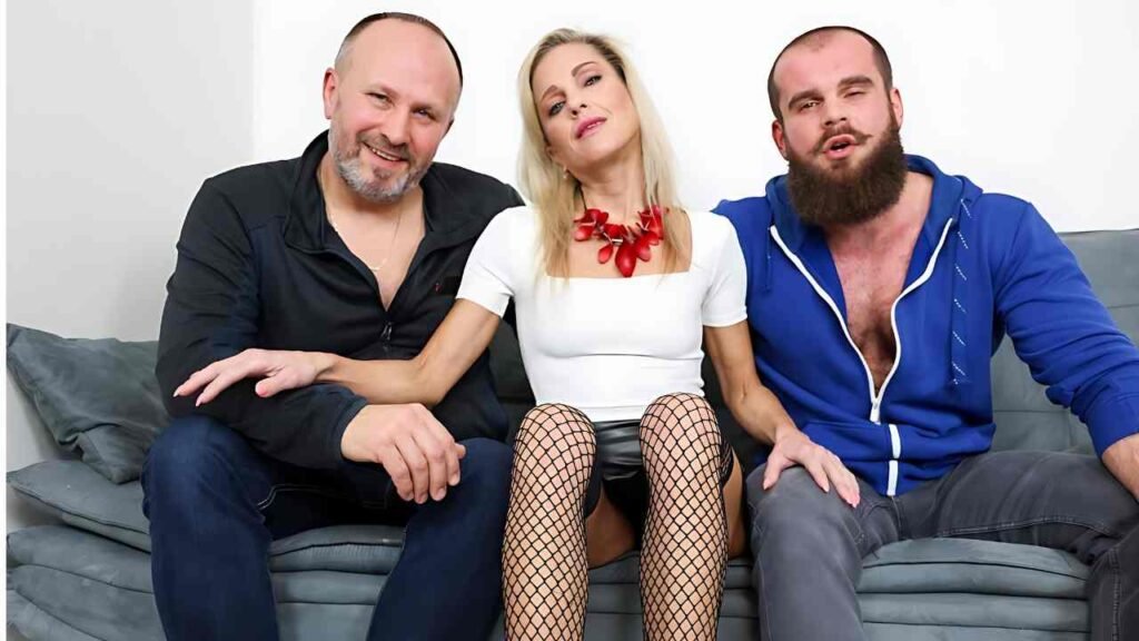 John Barber sit on a gray sofa: left man in black, center blonde in white top and red necklace, right bearded man in hoodie.