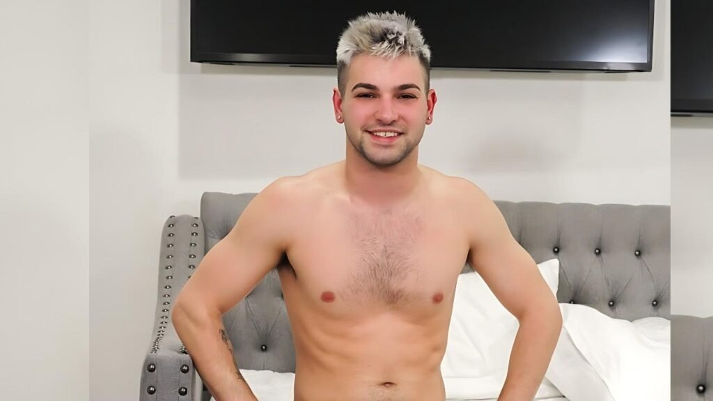 Johnny Rapid with short bleached hair sits on a grey tufted bed, hands on hips, in a bright room with white pillows.