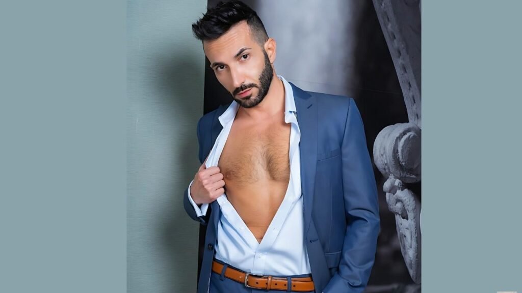 Johnny Viper in a blue suit with an open white shirt revealing chest hair, brown belt, gazing at the camera against a backdrop