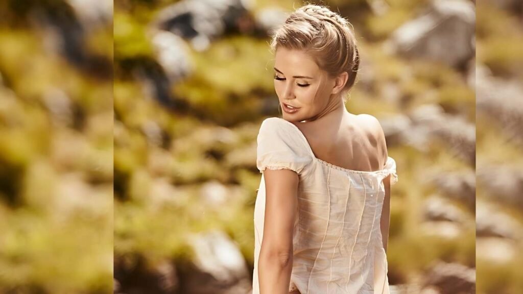 Julia Prokopy wearing a cream off-shoulder top, back to camera, glances over her shoulder in a warm, sunlit outdoor setting.