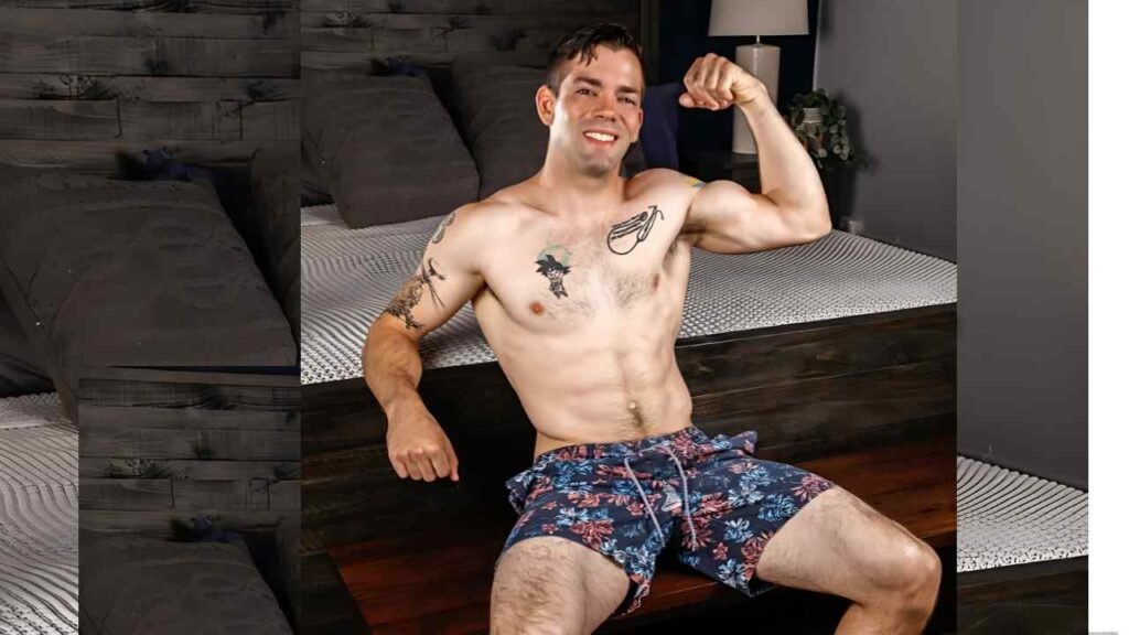 Julian Brady sits on a dark wood bed, flexing his right bicep; floral blue shorts and visible tattoos.