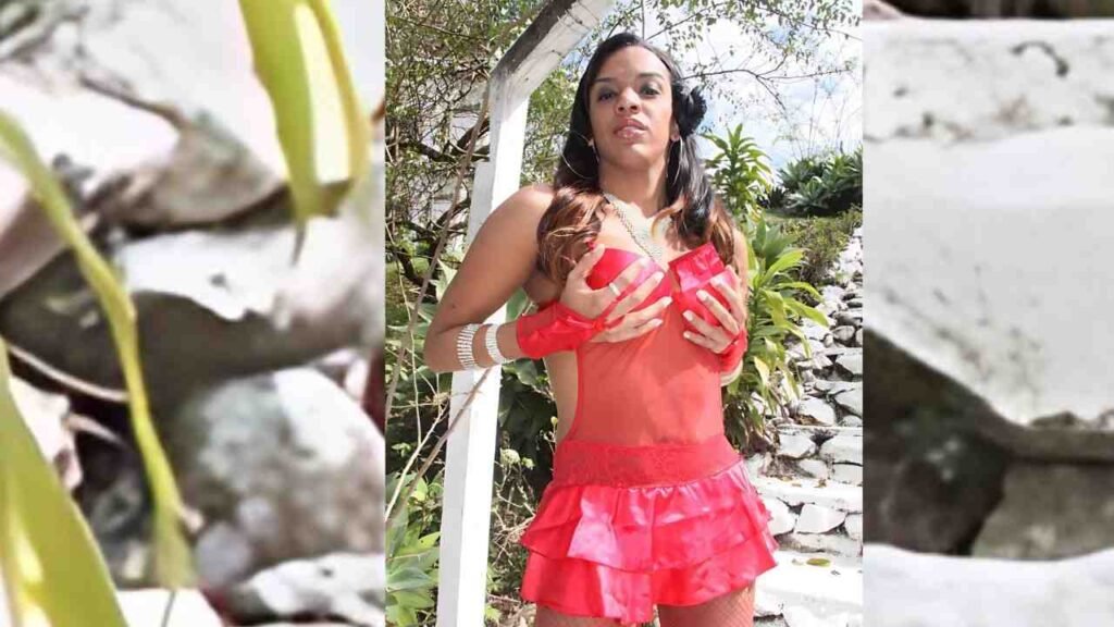 Kamilly Santos in a bright red ruffled dress and red gloves poses outdoors by a white railing, touching her chest.
