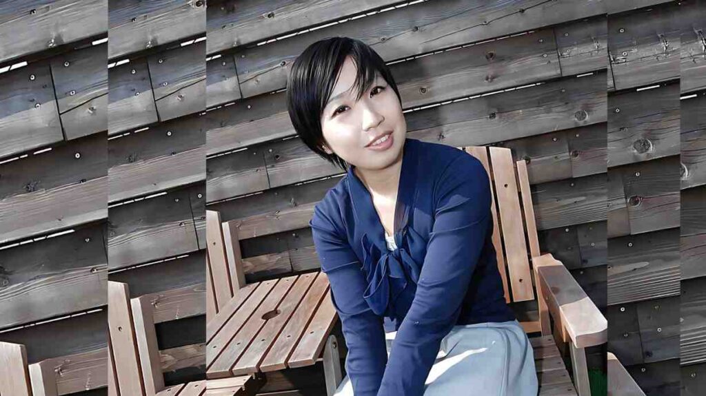 Kanako Kimura (木村夏菜子) in a navy blouse with a bow and light blue skirt sits on a wooden bench against a weathered wood wall.