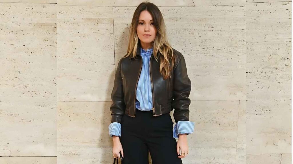 Kara Rose stands against a pale stone wall, wearing a brown leather bomber jacket over a blue striped shirt and black pants.