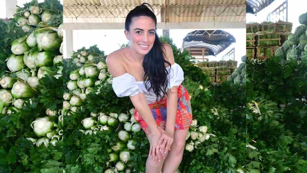 Karol Higuita in white off-shoulder top and colorful shorts bends forward among piles of green vegetables at a market.