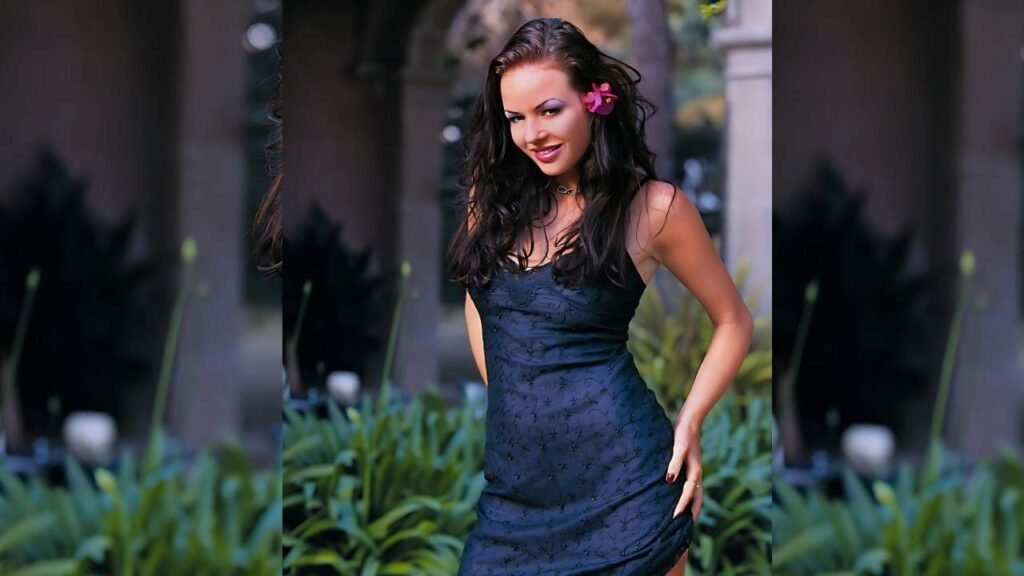Kate More in a dark blue lace dress stands among greenery, pink flower tucked behind her dark hair, smiling softly.
