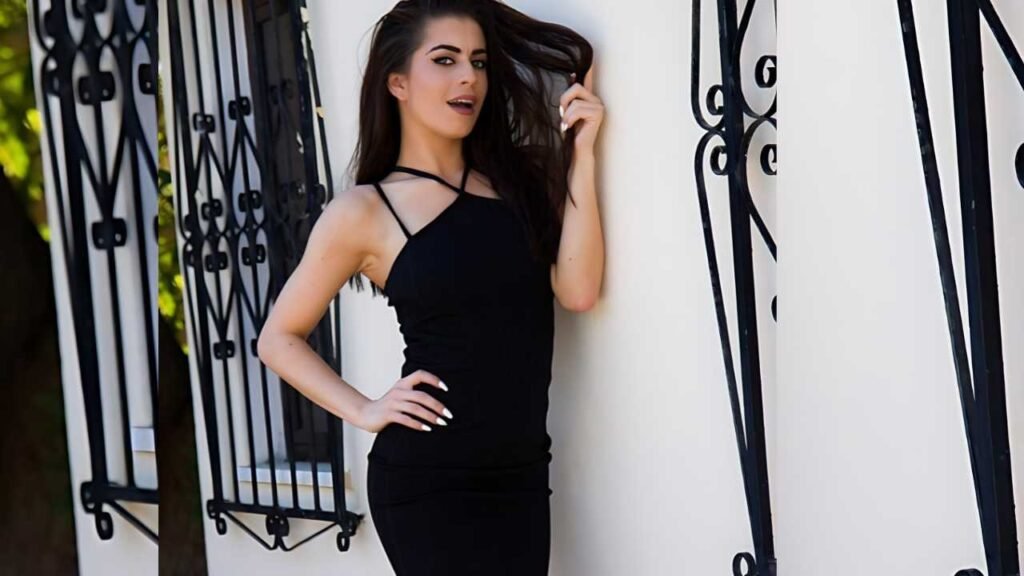 Katie Salmon in a black halter dress leans against a white wall beside ornate wrought-iron bars, hand on hip.