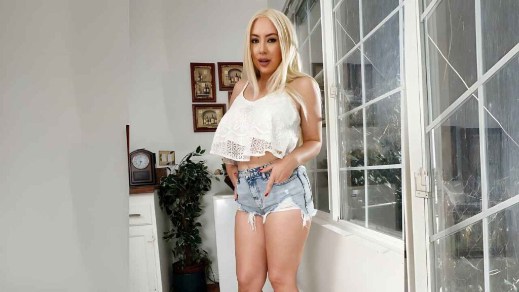 Kelly Paige in a white lace crop top and distressed denim shorts stands indoors by a large grid window, hands on hips.