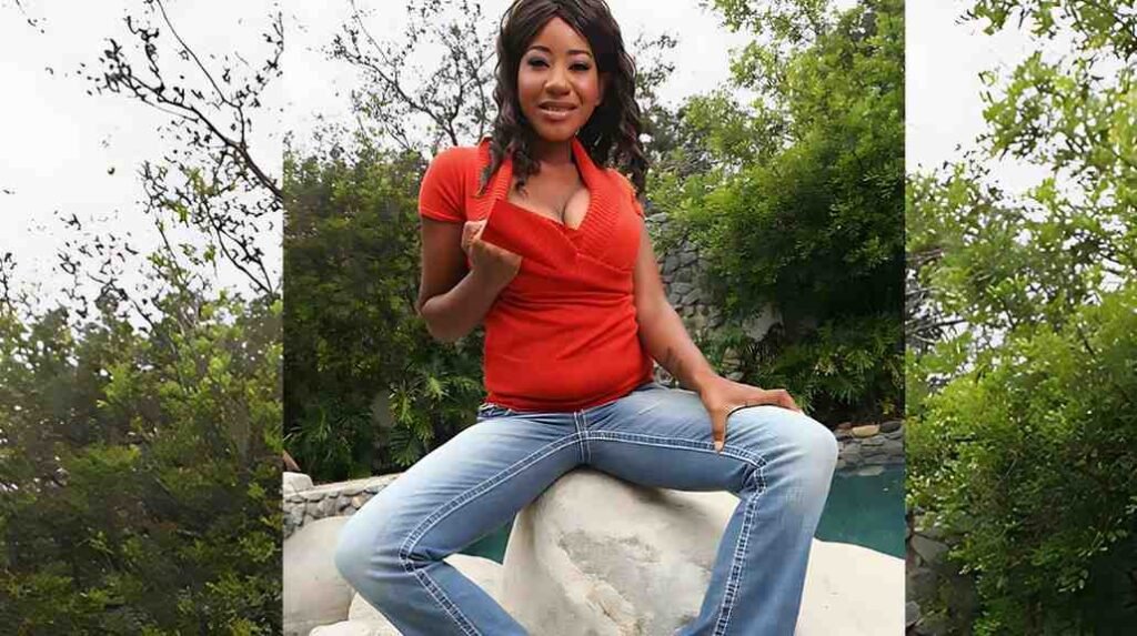 Kendra Lee in a red V-neck top and light jeans sits on a white rock by a pool, smiling with a lush green garden behind.