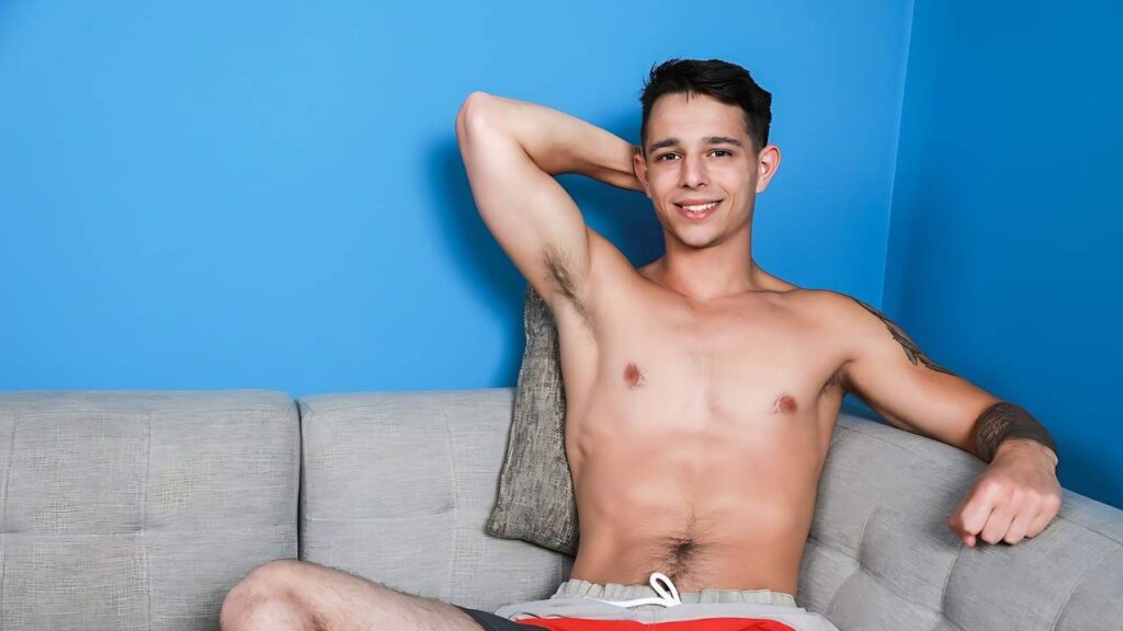 Kyle Wyncrest lounges on a gray couch against a bright blue wall, arm behind his head, wearing red shorts.