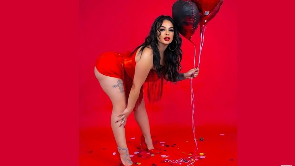 La Reina in a red fringe dress bends forward on a backdrop, holding red and black heart balloons; tattoo on thigh, hoop earring