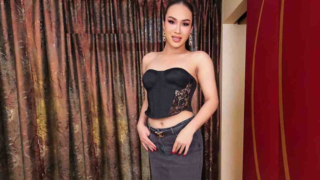Ladyboy Cindy in a strapless black corset with lace panels and a gray skirt, gold earrings, posing against brown curtains.