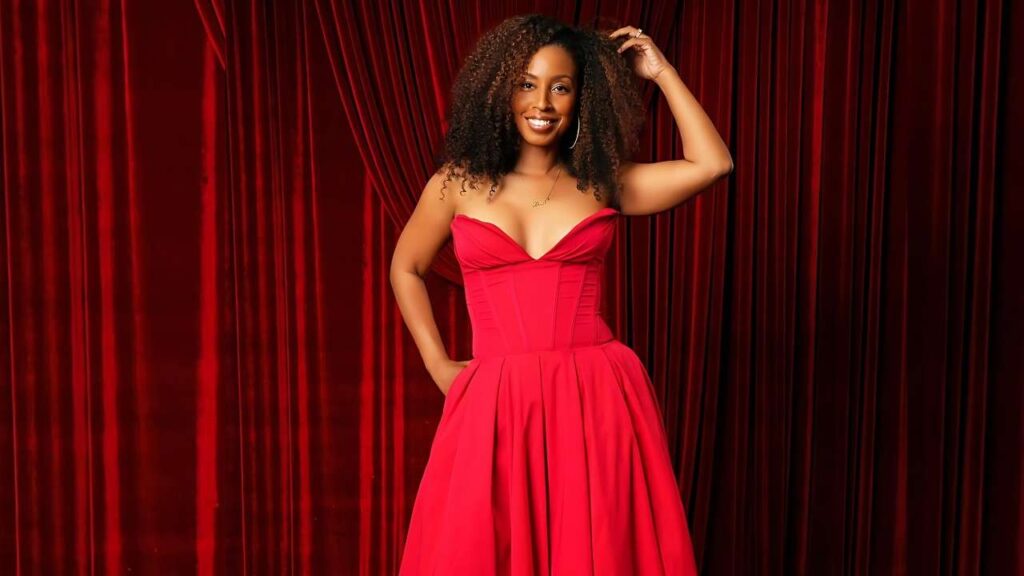 Lena Lewis in a red strapless corset dress poses before velvet curtains, one hand on hip, other touching her curly hair.