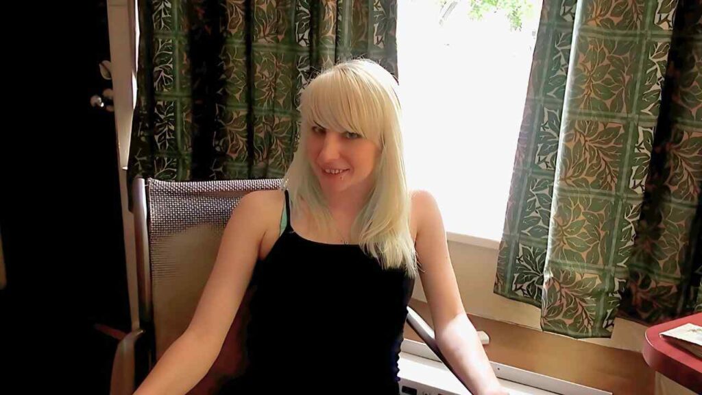 Lexi Jaxson in a black, thin-strap top sits on a chair by a sunlit window, green patterned curtains behind.