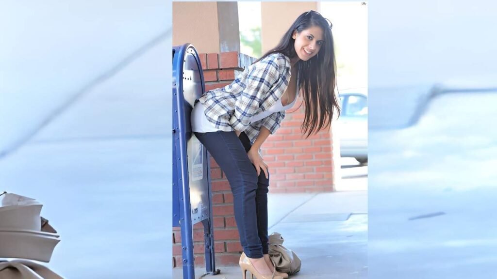 Lilly Anderson with long dark hair, plaid shirt over a white tank, dark jeans and nude heels, leaning on a blue mailbox beside brick pillars, smiling
