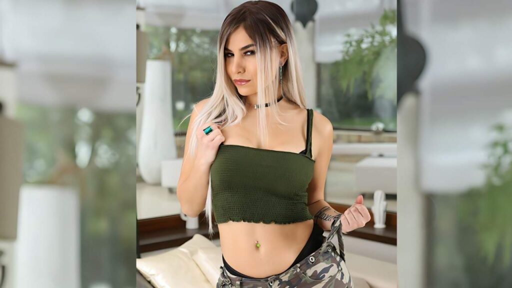 Lisa Gali with long blonde hair wearing a green cropped top and camo pants indoors, posing with strap and belly button piercing.