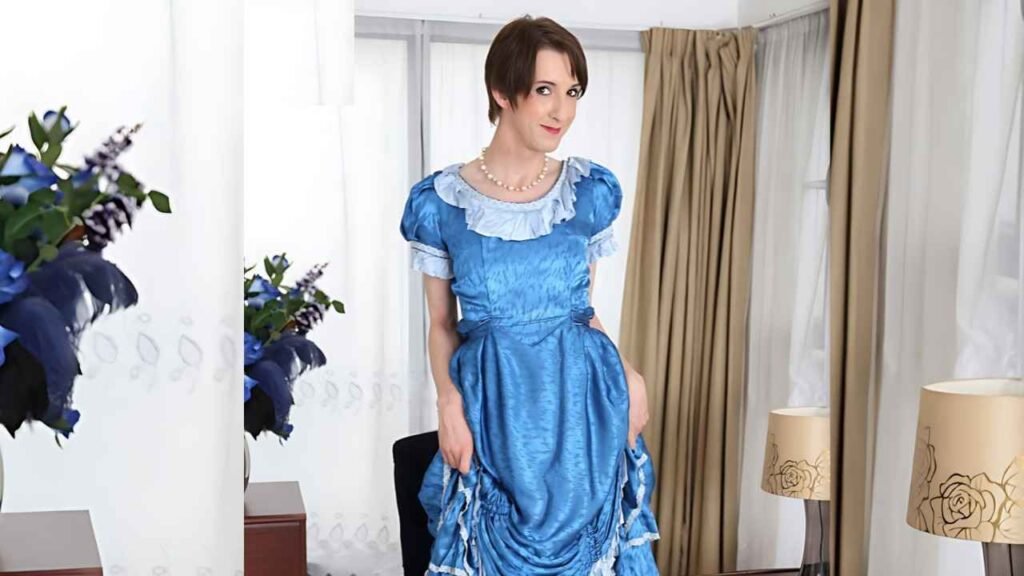 Lisa Heart in blue satin dress with ruffled neckline and puff sleeves, pearl necklace, stands beside beige curtains in a room.