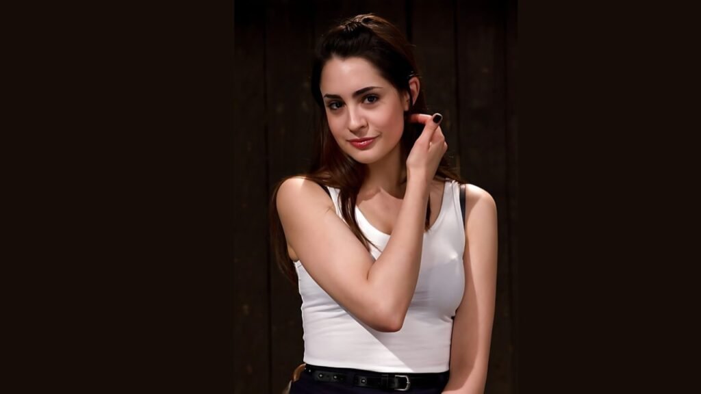 Lori Adorable wearing a white top poses confidently for the camera, smiling and looking directly at the viewer.