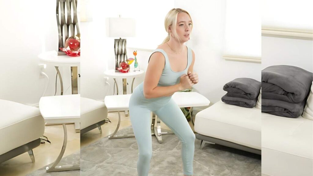 Lou Cipher in pale blue workout wear squats in a bright, modern white living room with a white sofa and grey blankets.