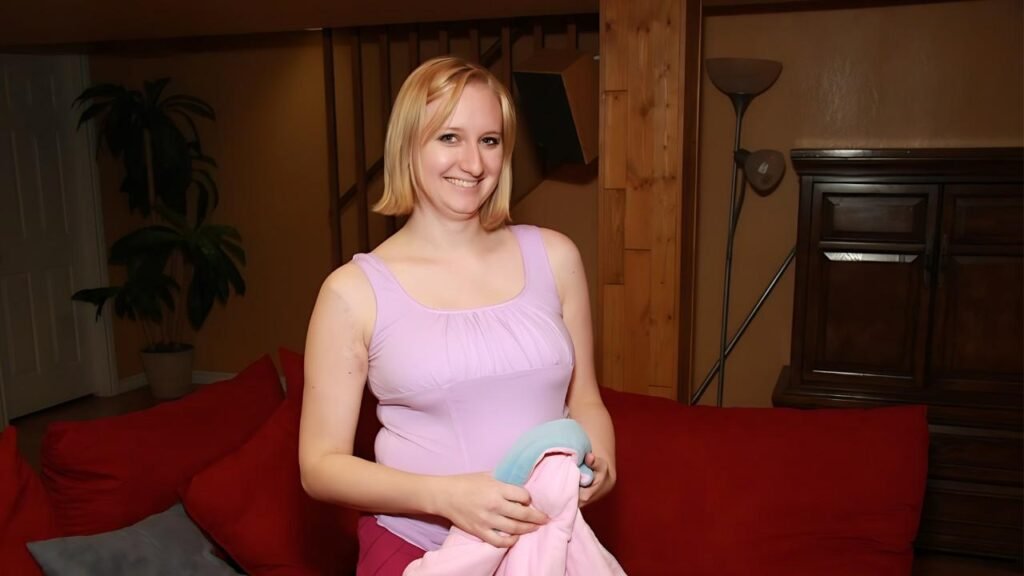 Maggie Star in a lavender tank top stands in a cozy living room, holding a pastel pink and blue blanket.