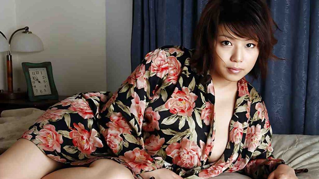 Mai Haruna on a bed in a floral robe with pink blooms; blue curtains behind and a vintage clock on the nightstand.