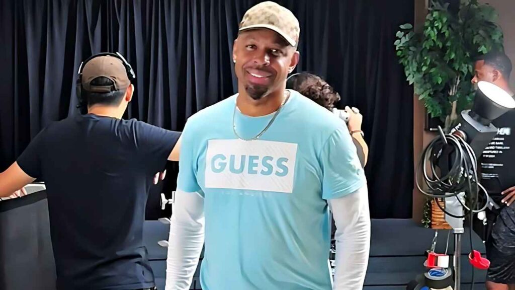 Marc Williams in a light blue GUESS shirt with white sleeves and a beige cap stands center in a studio, with crew and gear.