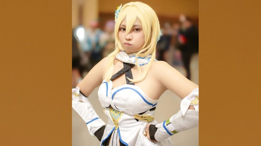 Mei Naomi with long blonde wig in white and blue armor, hands on hips, gold belt, blue flower hair clip, blurred convention crowd in background.