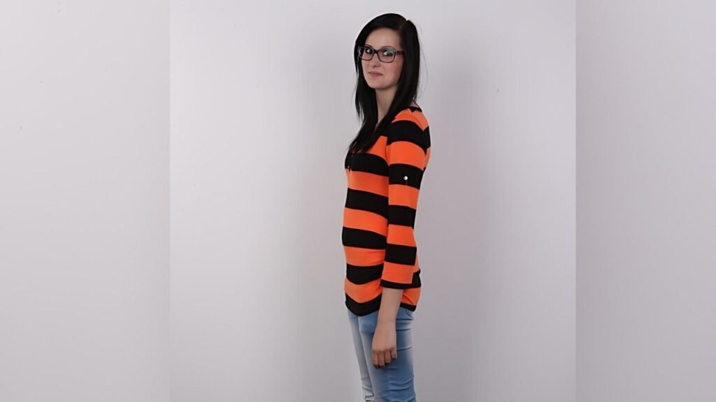 Mia Sky with hair and glasses, wearing orange and black striped sweater and blue jeans, against a gray wall.