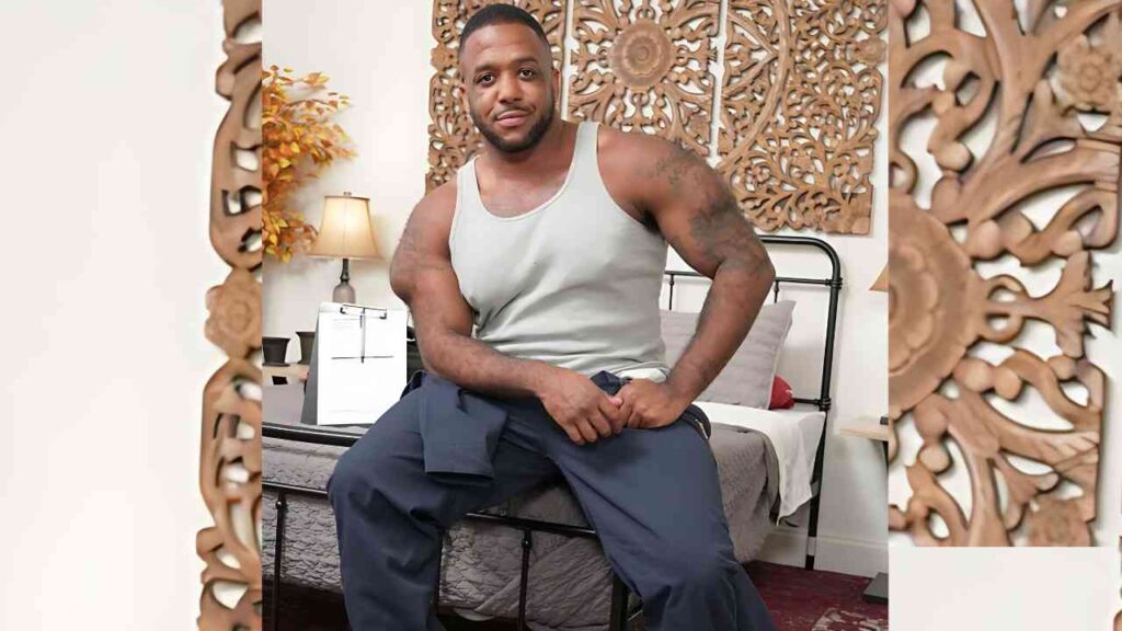 Micah Martinez in a gray tank top sits on a bed with blue pants, hands clasped, jacket draped over his lap, in a bedroom with ornate wood carvings and a lamp on the nightstand.