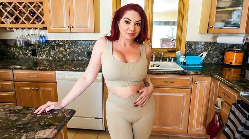 Mike Oxlong in a beige athletic set leans on a granite kitchen counter, smiling, with wooden cabinets and a sink behind.