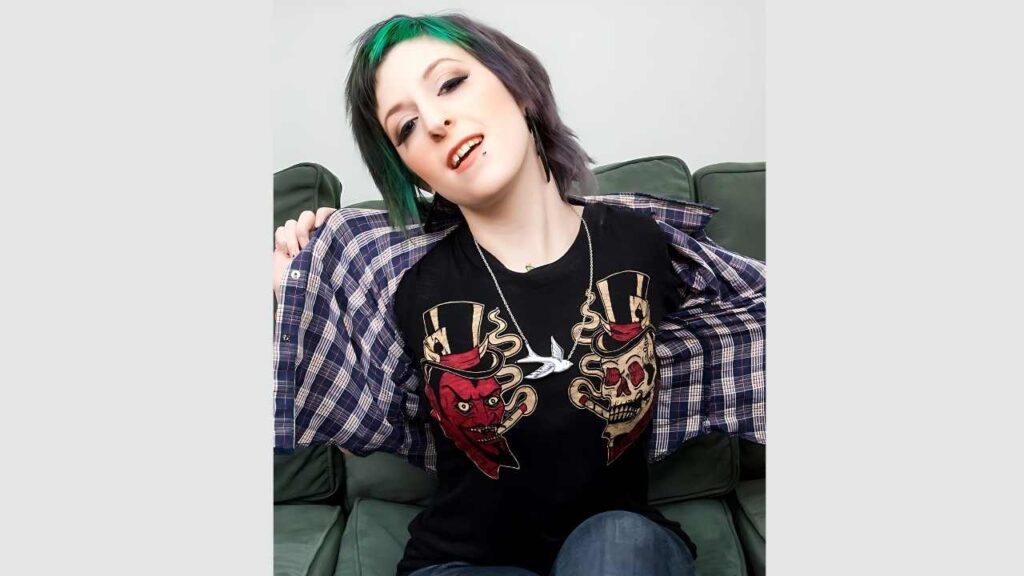 Miss Genocide with short black hair sits on a green couch, smiling, wearing a black tee with skulls and top hats, plaid shirt open
