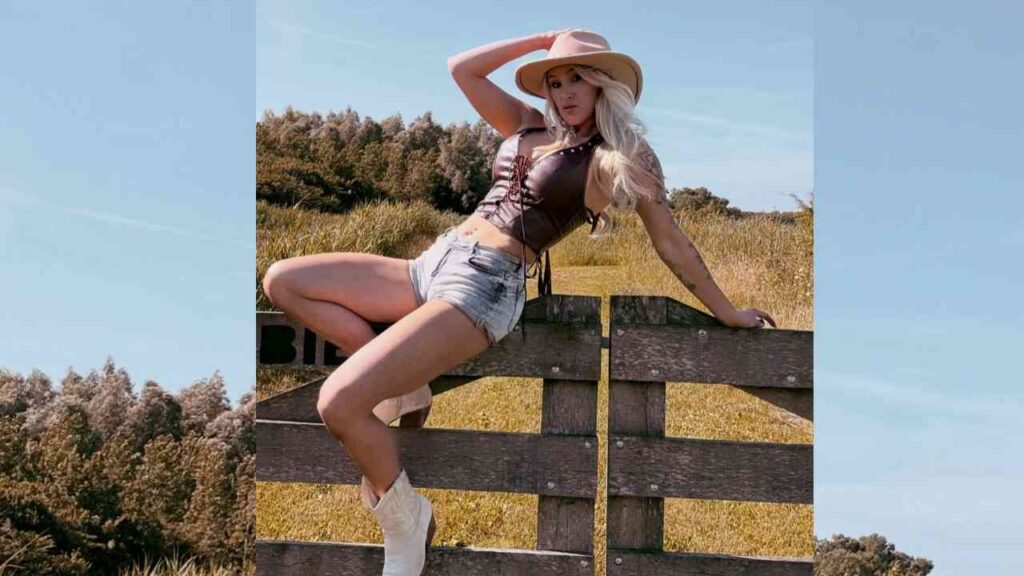 Miss Juicy Jane in a wide-brim hat, leather corset top, and cut-off denim shorts perches on a weathered wooden fence in a sunny rural field.