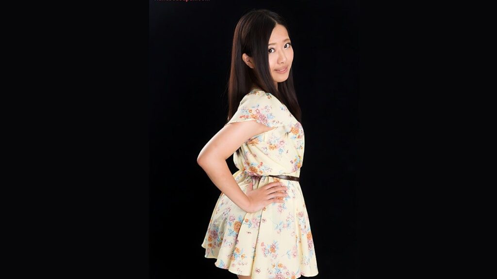 Miyuki Fukatsu in a pale yellow floral dress with a brown belt stands in profile against a black backdrop, smiling softly.