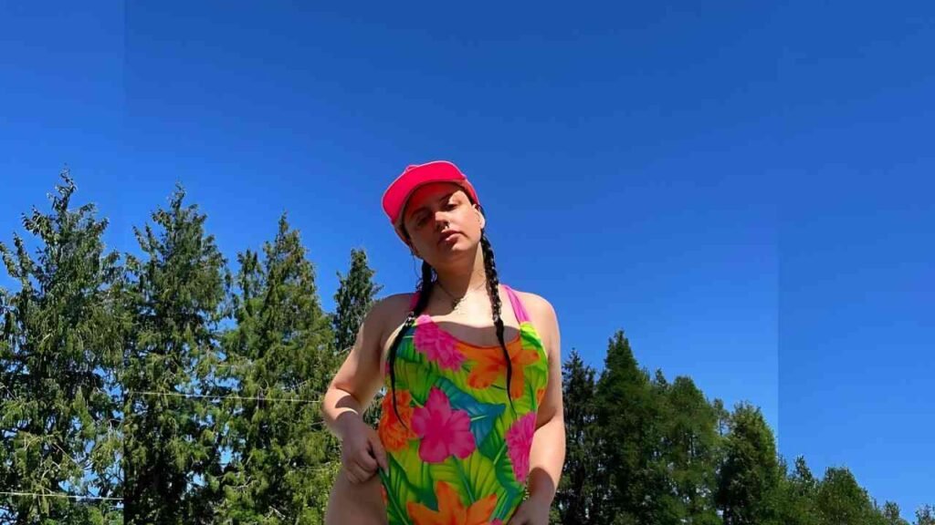 Molly Rae person outdoors under a clear blue sky, wearing a bright tropical swimsuit, pink cap, and two braids, with green trees behind