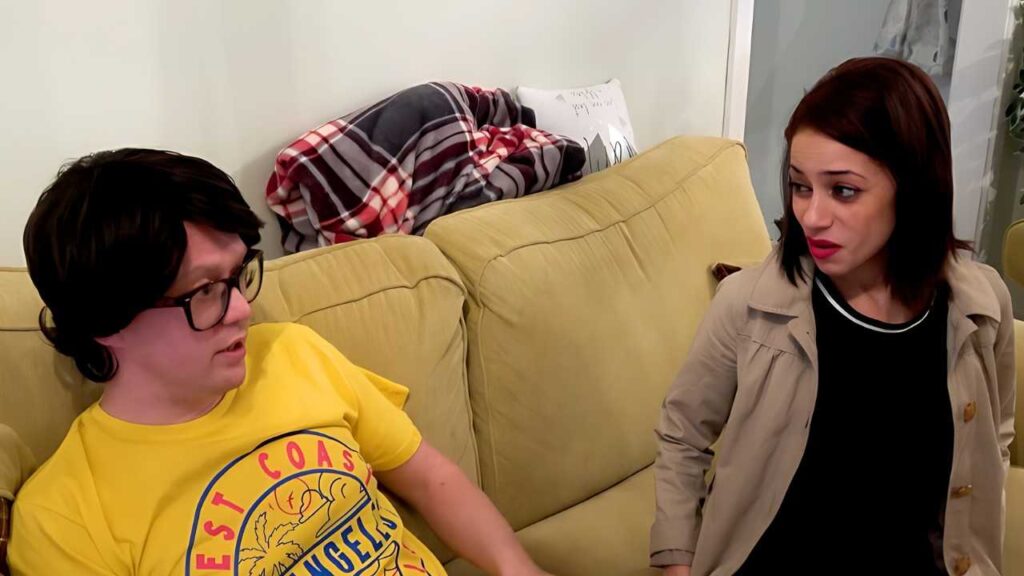 Myla Elyse sit on a beige couch, facing each other; one wears a yellow graphic tee and glasses, the other a tan trench coat.