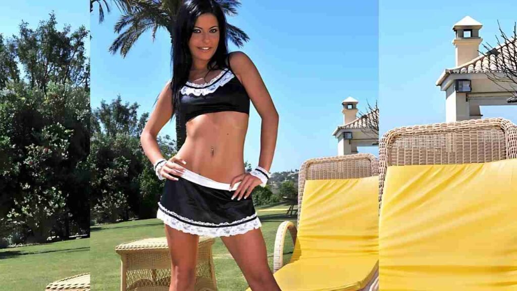Natalie Colt in a black and white maid outfit poses with hands on hips beside yellow-cushioned wicker chairs on a sunny lawn