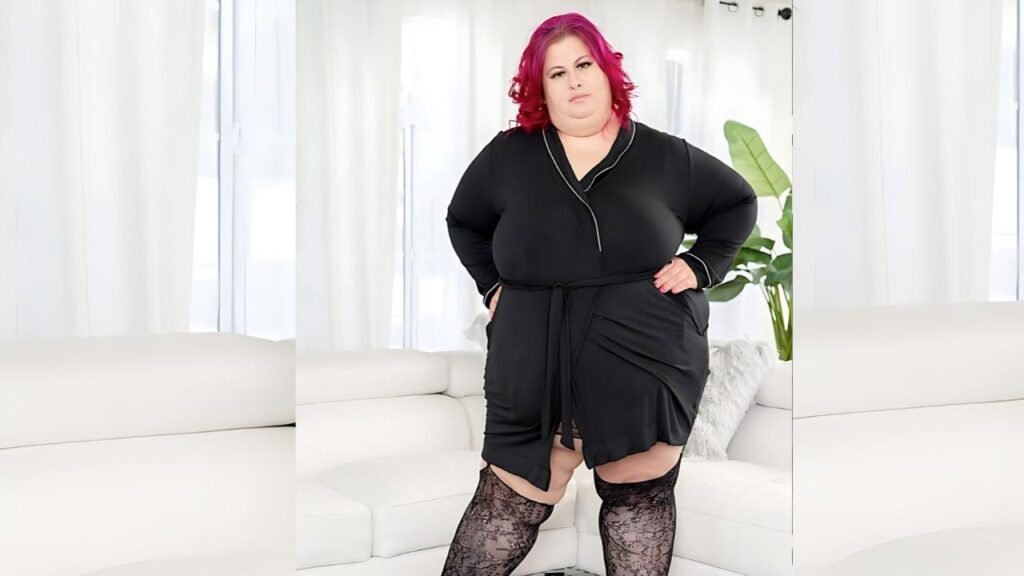 Natalie Kinky with pink hair in a black wrap dress, hands on hips, lace stockings, bright white living room.