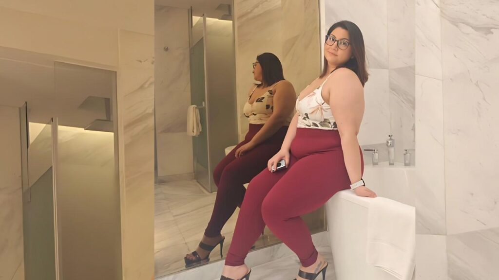 Nicole Lima with glasses sits on the edge of a white bathtub in a marble bathroom, wearing a floral top and burgundy leggings, reflected in a mirror